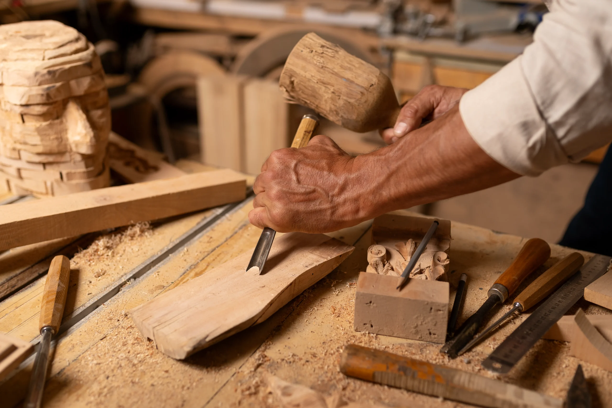 Close-up of hands using a mallet and chisel to carve wood, with various other carving tools and wood pieces on a workbench.