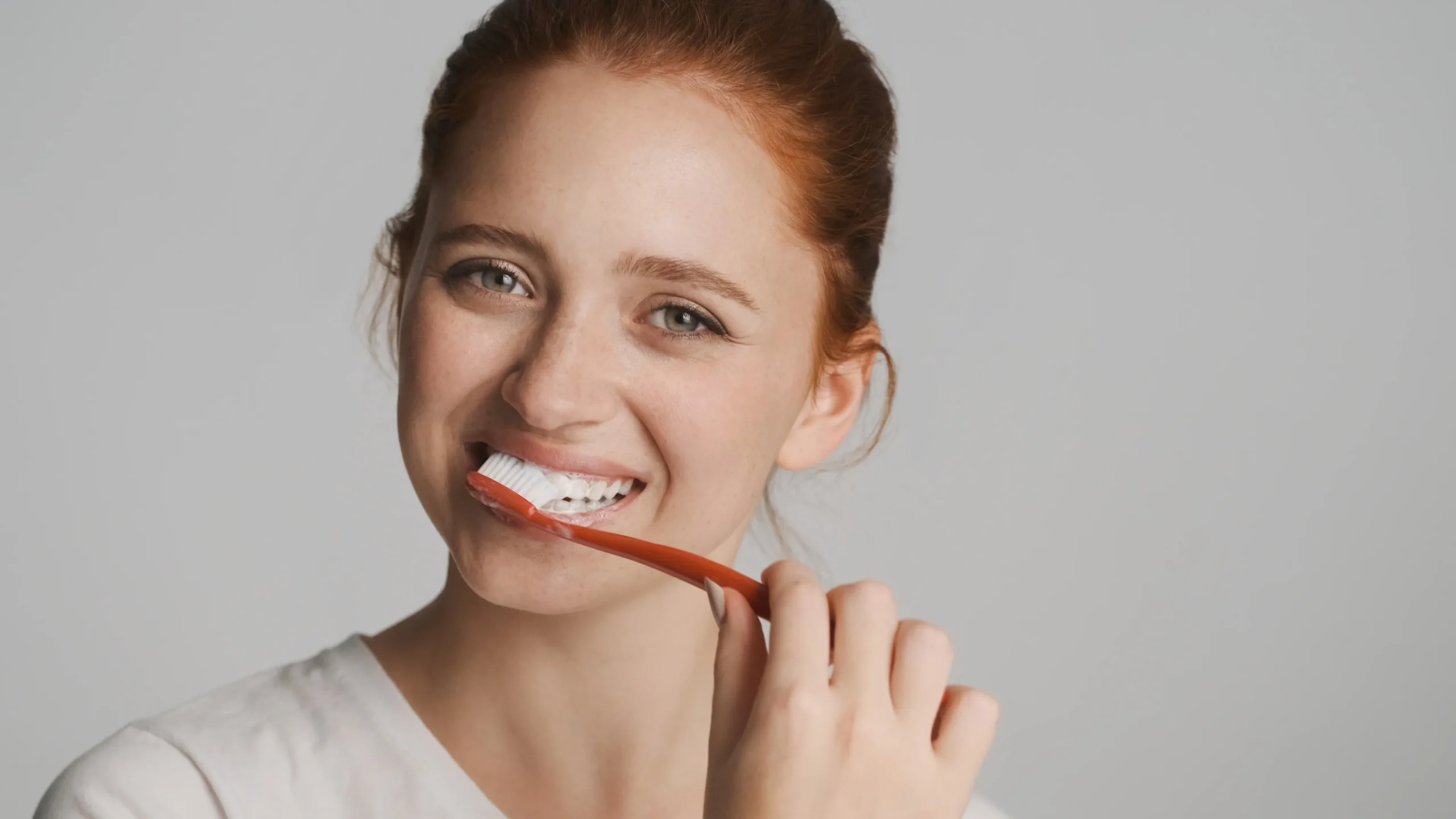 Smiling red-haired woman brushing her teeth with an orange toothbrush and white toothpaste, emphasizing the importance of oral hygiene for the health of teeth and gums, one of the benefits of ProvaDent.