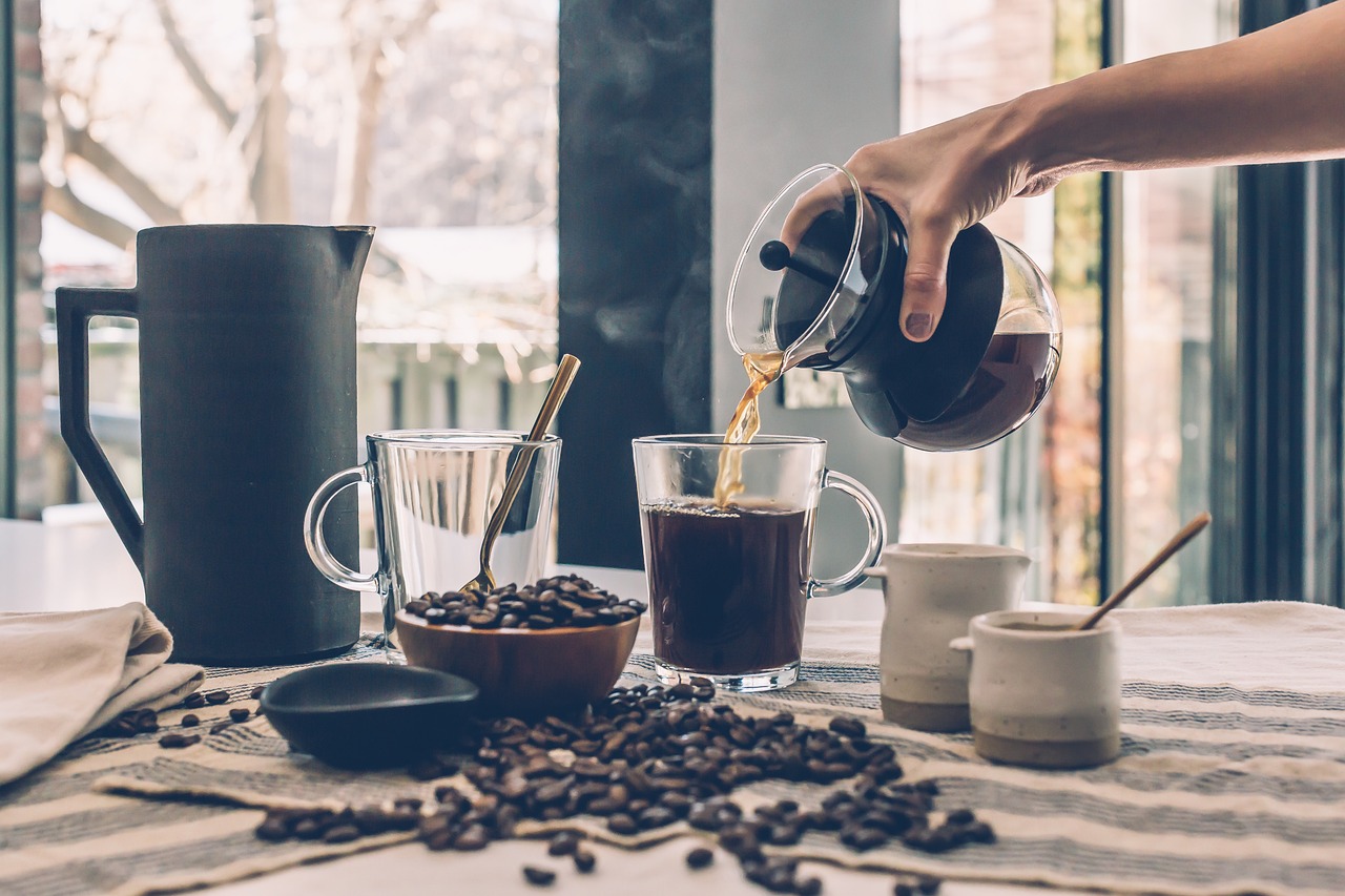 A hand pouring coffee from a French press into a cup, with coffee grounds scattered across the table, representing the preparation of the coffee for adding Java Burn.