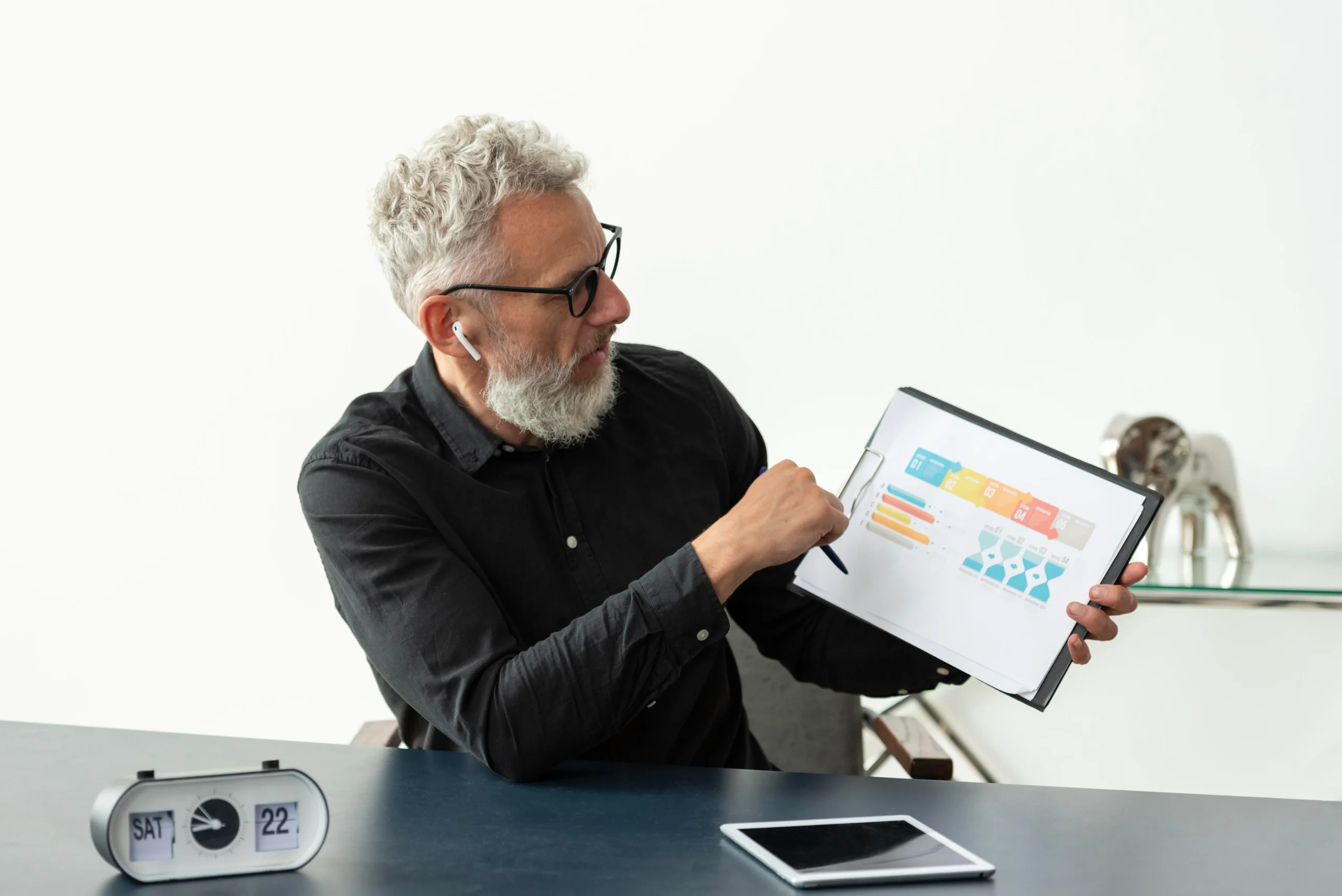 A mature man with gray hair and glasses, wearing earbuds, presents colorful charts on a clipboard, with a digital clock and tablet on the desk. The text overlay reads: "A Growing Demand for Innovative Solutions. As the number of older adults escalates, so does the need for innovative solutions to meet their healthcare and independent living requirements. In 2015 alone, an estimated 66 million Americans provided care to the ill, disabled, and elderly. By 2030, about 15 million Americans were utilizing some form of long-term care, including adult day care, home health services, nursing homes, and hospice care. However, navigating these various levels of care can be complex, with over 25% of nursing home residents facing hospital transfers each year."